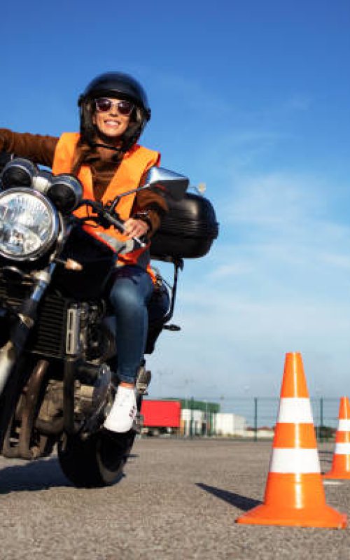 Female student with helmet taking motorcycle lessons and practicing ride. In background traffic cones and instructor with checklist rating and evaluating the ride. Motorcycle school of driving.